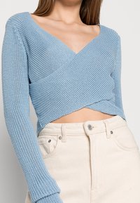 Maglione lavorato a maglia blu con un design avvolgente, maniche lunghe e scollatura a V. Tessuto testurizzato abbinato a pantaloni aderenti di colore chiaro.