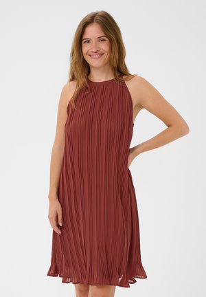 Femme souriante aux longs cheveux porte une robe plissée sans manches couleur rouille, posant avec une main sur la hanche devant un fond uni.