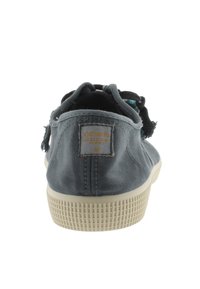 Sneaker da tela grigia con tessuto strutturato, suola in gomma e etichetta con logo sul retro. Presenta un design casual e lacci ad anello.