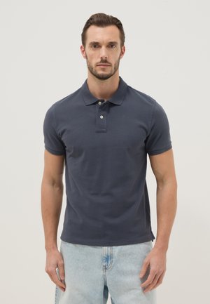 Hombre de pie mirando hacia adelante, vistiendo una camiseta tipo polo azul marina de manga corta y vaqueros azul claro contra un fondo liso.