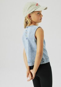 Top senza maniche in denim azzurro chiaro con scollatura rotonda, chiusura con bottoni sul retro e orlo grezzo. Abbinato a leggings neri e un cappellino chiaro.