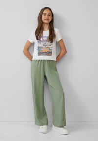 Weißes T-Shirt mit einem bunten Grafikdesign, kombiniert mit hellgrünen, lockeren Hosen. Das Model trägt weiße Sneakers.
