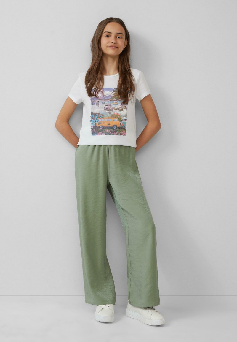 Weißes T-Shirt mit einem bunten Grafikdesign, kombiniert mit hellgrünen, lockeren Hosen. Das Model trägt weiße Sneakers.
