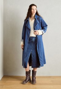Langer blauer Denim-Trenchcoat mit ausgeprägtem Kragen, cremefarbener Pullover darunter, Denim-Rock mit einem Schlitz vorne und braunen Stiefeln bis zur Mitte der Wade.