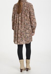 Tunique longue à manches longues à motif floral en rose pâle avec un col smocké, associée à des leggings noirs et des bottines beige.