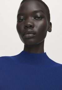 Modro rebrast džemper iz pletenine z visokim ovratnikom, ki prikazuje gladko teksturo. Tkanina ima strukturiran, oprijet dizajn. Barva kože je bogata in temna.