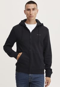 Svart zip-up hettegenser laget av mykt stoff, med en frontlomme, snorhette og subtilt logo på brystet. Standard passform.