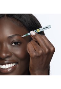 Kvinde med mørk hud bruger en Disney-tema TweezerMan pincet til at forme sin øjenbryn og smiler med hvide tænder mod en hvid baggrund.