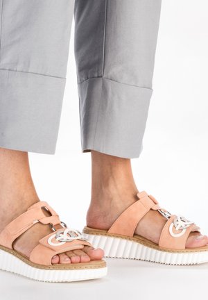 Sandales en daim rose avec des détails blancs, une boucle décorative et une semelle plate-forme texturée blanche. Portées avec un pantalon cropped gris.