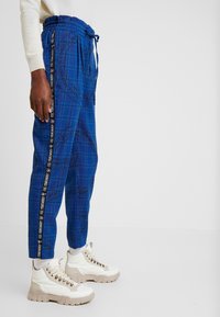 Blå rutiga joggers med elastisk midja, dragsko och svarta sidosträngar med text. Kombinerat med vita och beige höga sneakers.