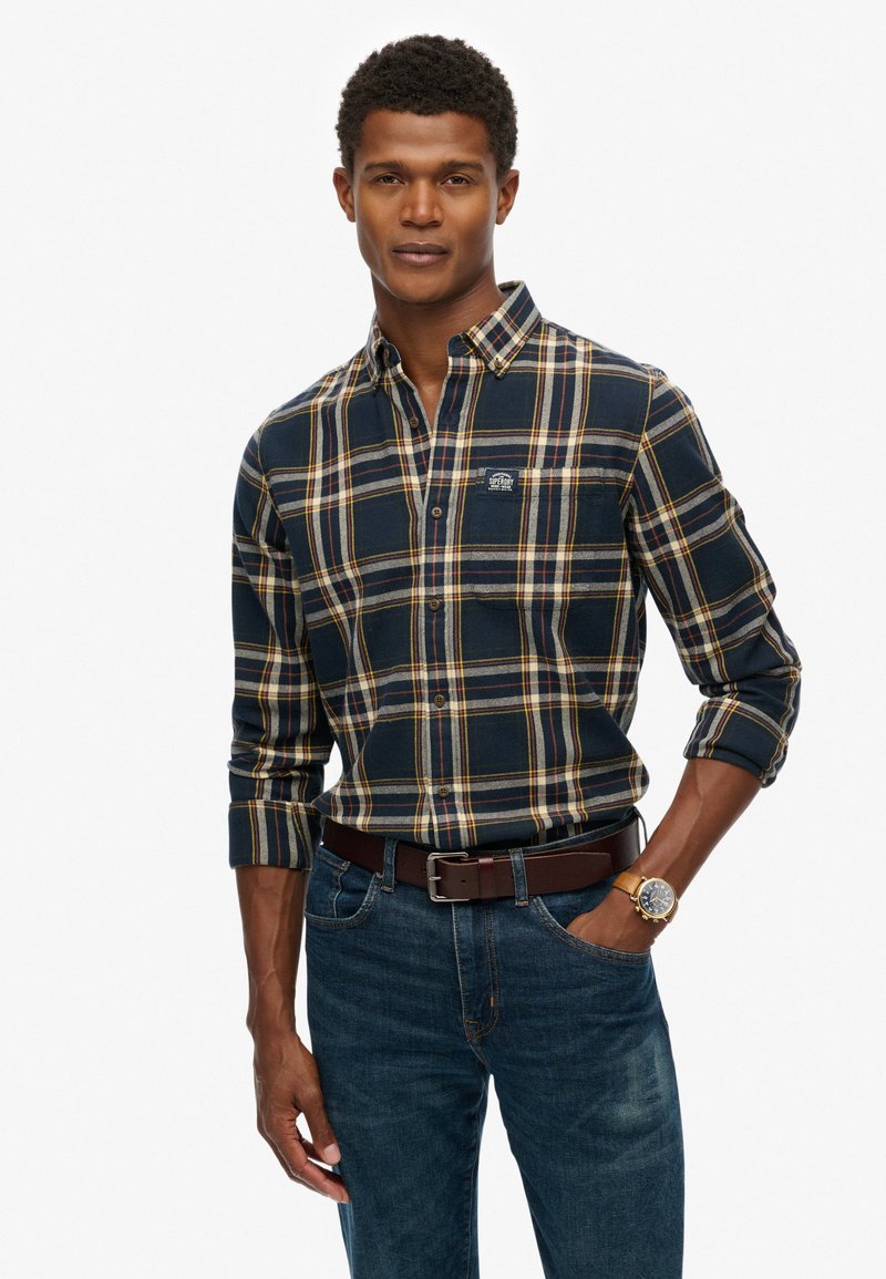 Chemise à carreaux bleu marine et jaune avec col boutonné, manches longues et un écusson, associée à un jean bleu et une ceinture brune.