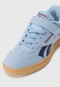 Light blue suede sneaker with lace-up design and a Velcro strap. Features dark blue accents and a gum rubber sole. Reebok logo on the tongue.