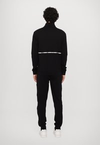 Black sweatshirt with high collar and reflective strip, paired with black sweatpants and white sneakers. Texture appears smooth and soft.