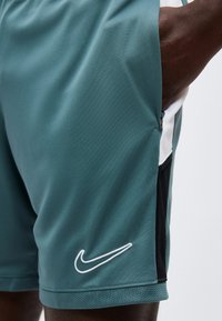 Κοντινό πλάνο προσώπου που φοράει μπλε-πράσινα αθλητικά σορτς με λευκό λογότυπο Nike και μαύρες πλαϊνές ταινίες, χέρι ακουμπά μέσα στην τσέπη με φερμουάρ.