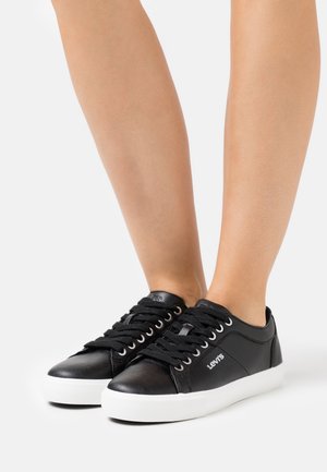 Baskets en cuir noir avec semelles en caoutchouc blanc, dotées d'un bout arrondi, d'un laçage classique à l'avant et d'un logo sur le côté.
