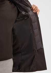Donkere jas met een zachte binnenvoering; heeft een ritsvak en een gewatteerd patroon aan de binnenkant. Gedetailleerde stitching langs de randen.