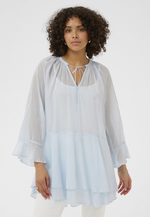 Vrouw met krullend haar, gekleed in een lichtblauwe doorschijnende blouse met lange mouwen, gerimpelde zoom en een strik bij de hals, gecombineerd met een witte broek.