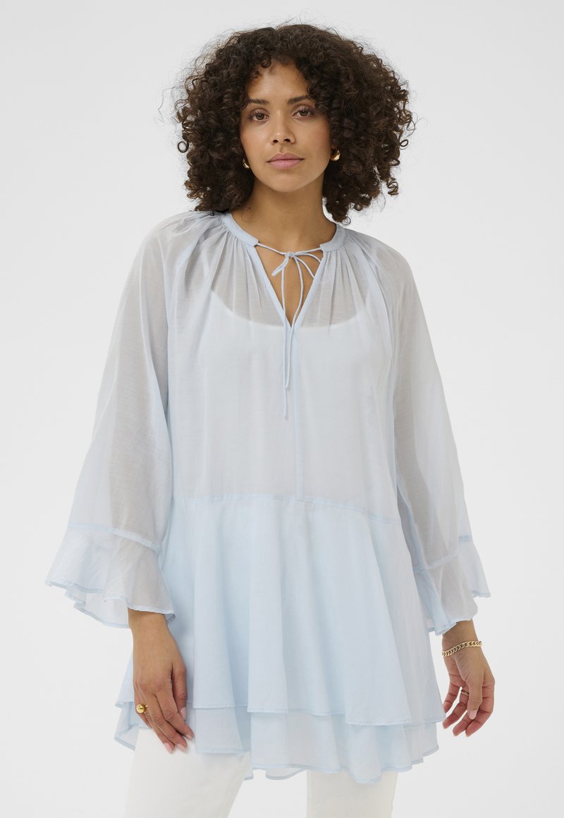 Femme aux cheveux bouclés portant un chemisier à manches longues en voile bleu clair avec ourlet à volants et lien au col, associé à un pantalon blanc.