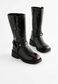 Schwarze knöchellange Stiefel aus glänzendem Leder, seitlichem Reißverschluss, verstellbarem Riemen mit Hardware und auffälliger Gummisohle.