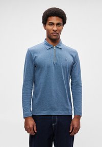 Sinine, pika varrukaga polosärk lukuga kaelusega ja peene logoga rinnal; valmistatud pehmest, tekstuurse kangaga.