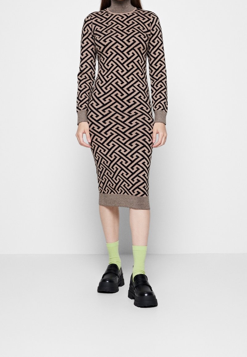 Femme portant une robe marron à manches longues arrivant au genou avec un motif géométrique noir, des chaussettes vert néon et des chaussures à semelles compensées noires, debout sur un sol blanc.