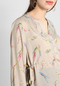 Helle beigefarbene Bluse mit Knopfleiste, die farbenfrohe Vogelmuster zeigt, lange Ärmel hat und eine geraffte Taille mit einem gebundenen Detail aufweist.