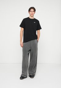 Camiseta de algodão preta com um pequeno logotipo branco, combinada com calças cinzentas largas em um tecido macio e texturizado. O modelo usa sapatos pretos.
