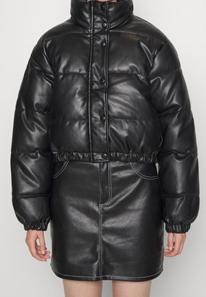 Woman wearing black cropped puffer jacket and black leather skirt with white stitching, standing against plain light background.