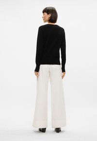 Maglione nero con maniche lunghe, scollatura rotonda e polsini a coste, abbinato a pantaloni bianchi a gamba larga e scarpe con tacco nere.