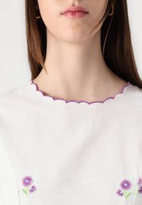 Blusa bianca con scollatura viola ondulata e fiori ricamati viola all'orlo. Tessuto morbido con dettagli di texture minimi.