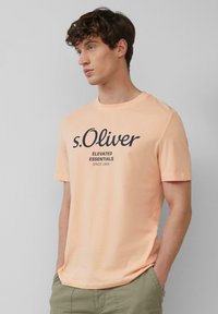 Perzikkleurig katoenen T-shirt met korte mouwen, ronde halslijn en donkerblauwe tekst die zegt "s.Oliver ELEVATED ESSENTIALS SINCE 1969."