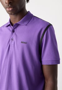 Μωβ πόλο μπλούζα με ριχτό γιακά, μαύρες πλευρικές λεπτομέρειες και κεντημένο λογότυπο "BOSS". Κατασκευασμένη από απαλό ύφασμα, διαθέτει κλείσιμο με δύο κουμπιά.