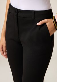 Gros plan sur une personne portant un pantalon noir ajusté, la main dans la poche et plusieurs bracelets au poignet.