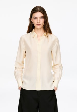 Jasnożółta koszula z długim rękawem wykonana z gładkiej, lekkiej tkaniny, zapinana na guziki z klasycznym kołnierzykiem, zestawiona z czarnymi spodniami.