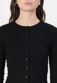 Čierny rebrovaný top s dlhým rukávom a okrúhlym výstrihom, s siedmimi čiernymi gombíkmi na prednej strane, ktorý má priliehavý strih a hladkú textúru.