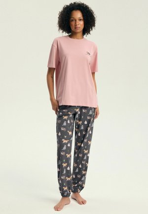 Kobieta stoi boso, ubrana w luźny różowy t-shirt i ciemne spodnie do piżamy z różnymi ilustracjami kotów na jasnym tle.