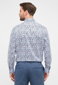 Camicia abbottonata con motivo floreale blu e nero su sfondo bianco, con maniche lunghe e colletto, abbinata a pantaloni blu.