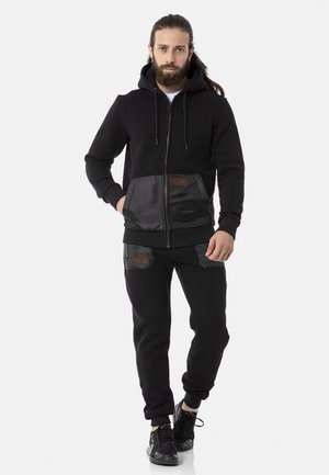 Schwarze Zip-Hoodie mit einer kontrastierenden glänzenden schwarzen Fronttasche; kombiniert mit passenden Jogginghosen, die über strukturierte Taschendetails und Bündchen verfügen.