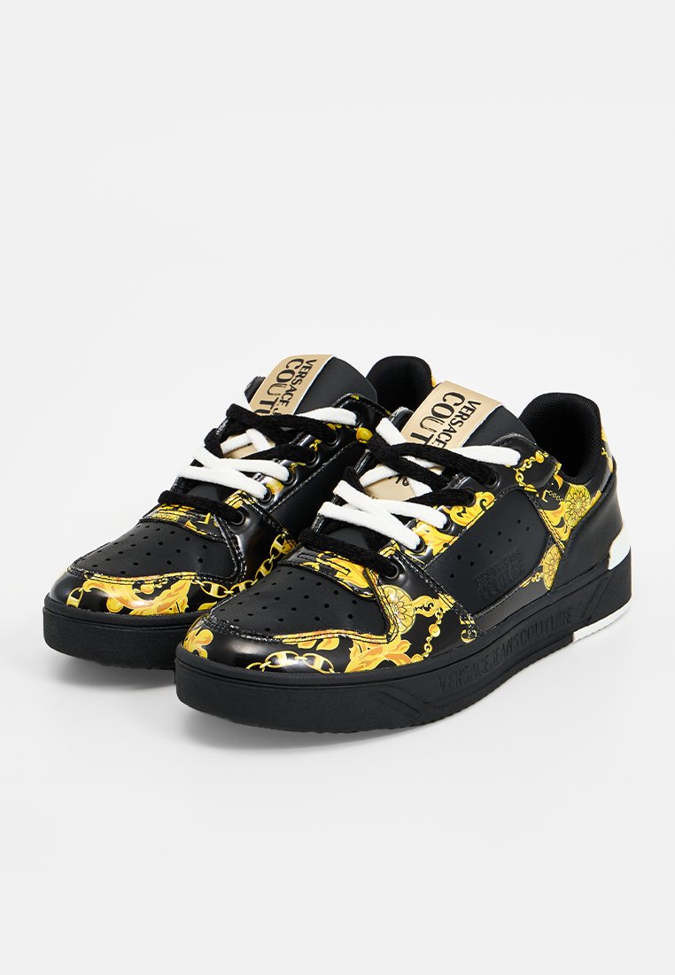 Baskets basses noires avec des motifs baroques ornés jaunes, lacets blancs et noirs, et étiquette "Versace Jeans Couture" sur la languette et la semelle.