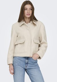 Ung kvinne med langt brunt hår iført en kremfarget ulljakke med klaffelommer og lyseblå høytlivsjeans, hånden i lomma.