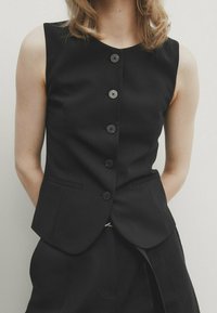 Schwarzes ärmelloses Hemd mit tailliertem Design, ausgestattet mit einer Reihe von sechs Knöpfen, zwei vorderen Taschen und einer glatten Textur.