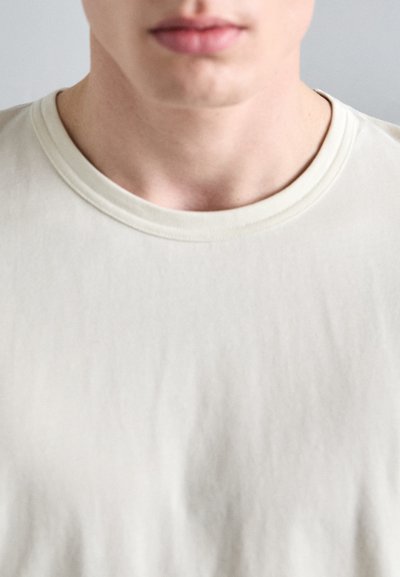 Tricou bej deschis, cu guler rotund și textură moale, prezentând un design minimalist, fără modele sau accente vizibile.