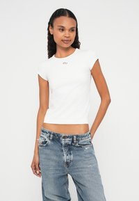 Balta, pieguļoša t-krekls ar īsām piedurknēm un apaļu izgriezumu. Uz krūtīm ir neliels metālisks logotipa elements.