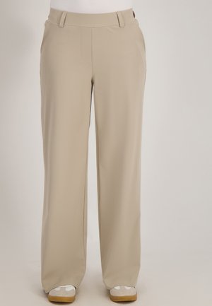 Beige recht gesneden broek met riemlussen, zijzakken en witte sneakers met gumzolen die onderaan zichtbaar zijn.