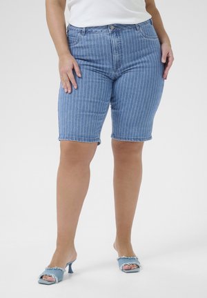 Kvinde iført blå stribede denimshorts, hvid top og lyseblå åbne hælede sandaler stående mod en ensfarvet baggrund.