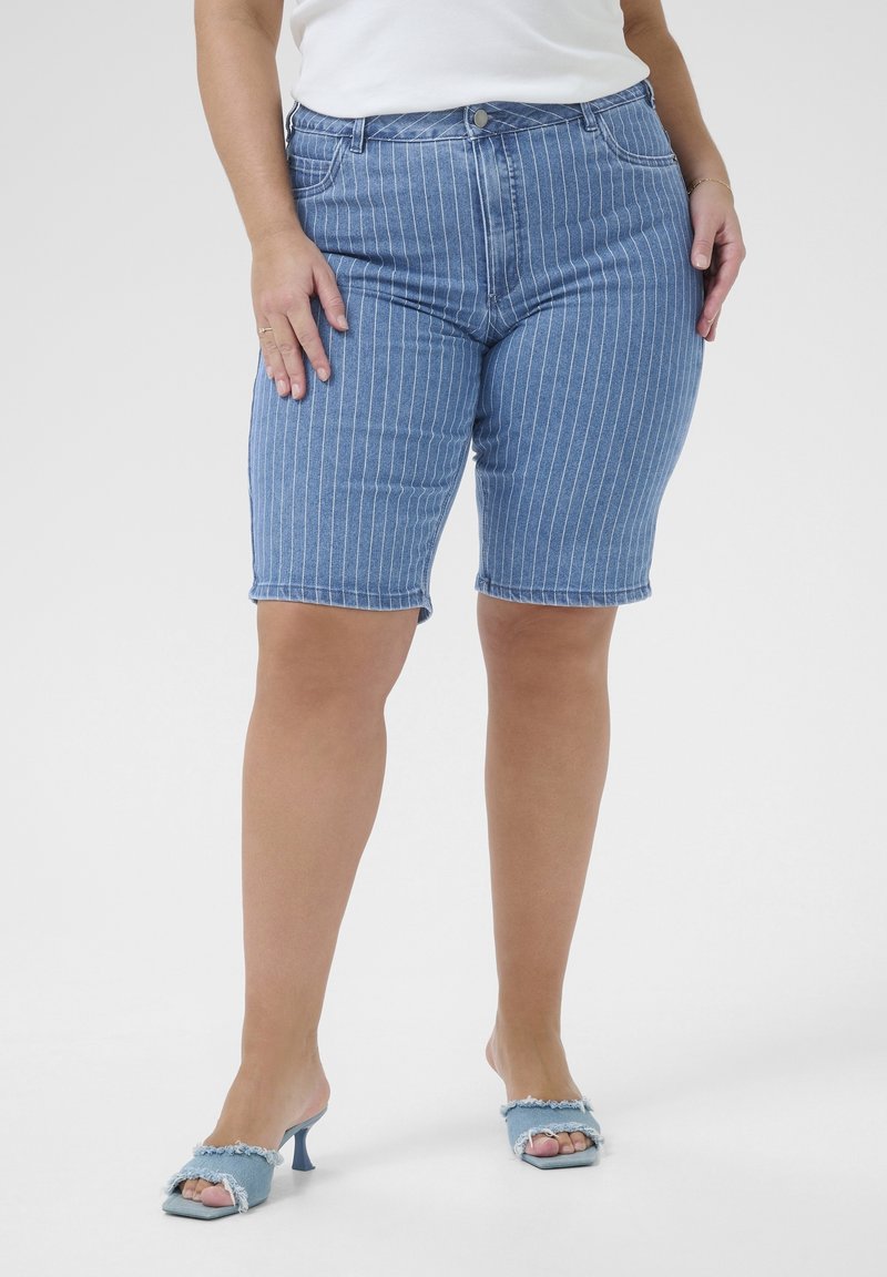 Femme portant un short en jean bleu à fines rayures, un haut blanc et des sandales à talons ouvertes bleu clair, debout devant un fond uni.