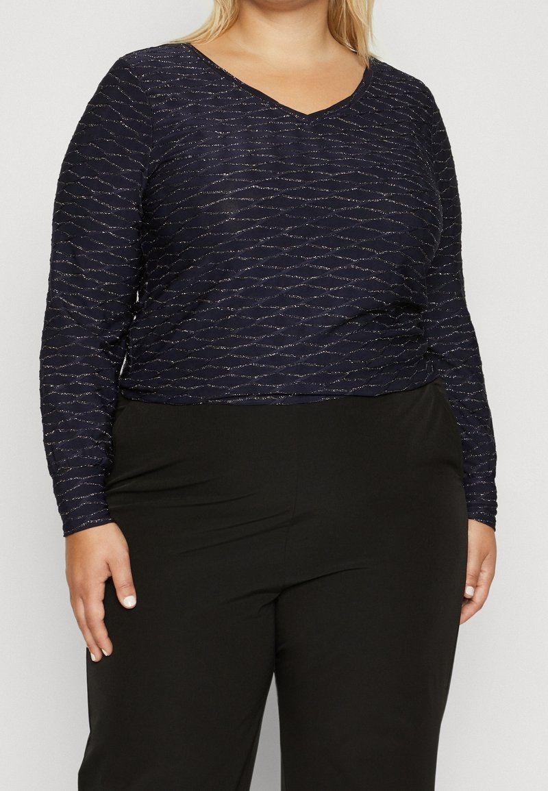 Marinblå långärmad topp med V-ringning, som har ett strukturmönster med vågor och metalliska detaljer. Matchad med svarta-byxor med hög midja.