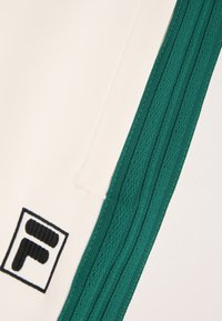 Pantalones cortos deportivos blancos con franjas elásticas laterales verdes y un parche de logo negro con una letra "F" estilizada. Textura de tela suave.