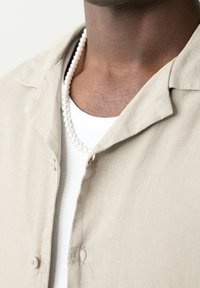 Gros plan d'une personne portant un t-shirt blanc à côtes, une chemise beige à boutons et un collier de perles blanches autour du cou.