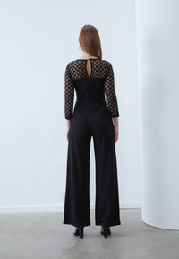 Mono negro con mangas largas de encaje semitransparente que presentan un patrón geométrico, piernas anchas y un alto respaldo con una abertura en forma de llave.